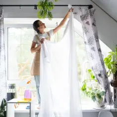 Kobieta w fartuchu wiesza białą firankę, przygotowując się do jej prania. W tle widać okno i rośliny.