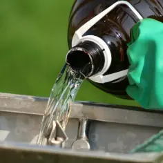 Ręka w zielonej rękawicy przelewa płyn z butelki. Woda h202 spływa do metalowego pojemnika.
