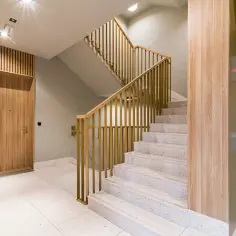 Nowoczesna klatka schodowa z drewnianymi panelami i złotymi balustradami. Praktyczne ściany na klatce schodowej dodają ciepła i elegancji. Obok winda.