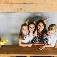 Dzieci w czapkach urodzinowych pozują przy drewnianej fotobudce z kartonu. Napis "Happy Birthday" zdobi górę.