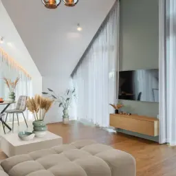 Przytulny salon z sofą w kolorze kremowym, stołem i telewizorem. Jasne zasłony dodają lekkości.